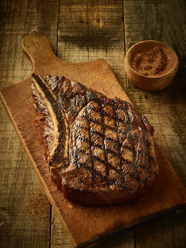 Large steak with bone on a wooden cutting board. Off to the side is a small bowl of seasonings.