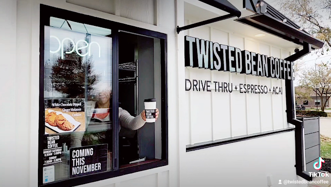 Exterior close view of a drive-thru window. The building has white slats and black accenting. The sign off to the right side of the window says "Twisted Bean Coffee, drive thru + espresso + acai." A hand holding a hot coffee cup peaks out of the drive-thru window.