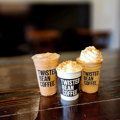 Three coffee set on a wooden table. The one on the left is an iced coffee, the one in the middle is a hot latter with whipped cream and caramel, and the one on the right is an iced coffee with whipped cream and caramel. All three say "Twisted Coffee Bean" on the front of the cups.