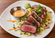 Pepper-crusted seared tuna sliced over a fresh shredded salad with green onions, wasabi, and a small cup of creamy dipping sauce on a white plate.