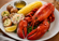Whole cooked lobster served with corn on the cob, baby potatoes, coleslaw, melted butter, and a lemon wedge on a white dinner plate.