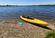 Yellow kayak resting on a pebbled lakeshore beside a teal paddle with calm blue water and a tree-lined shoreline in the background.