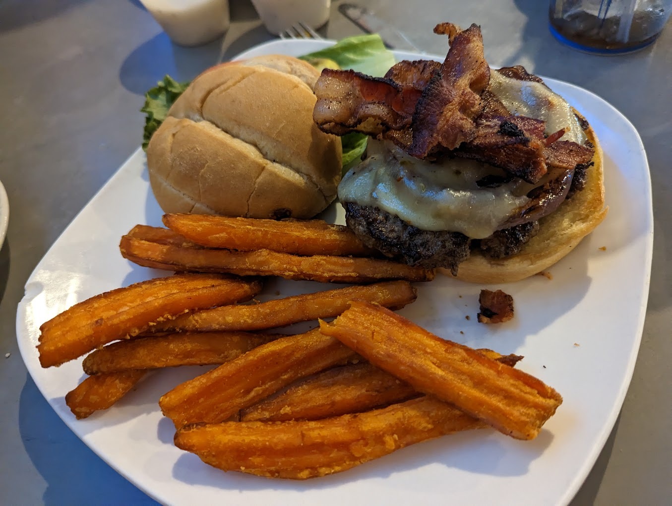 A white plate with sweet potato fries and a burger with cheese and bacon.