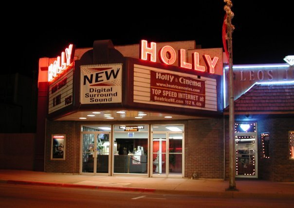 An outside nighttime view of a movie theater entrance with a large white letterboard sign awning out front, and large red lighted lettering reading "Holly"