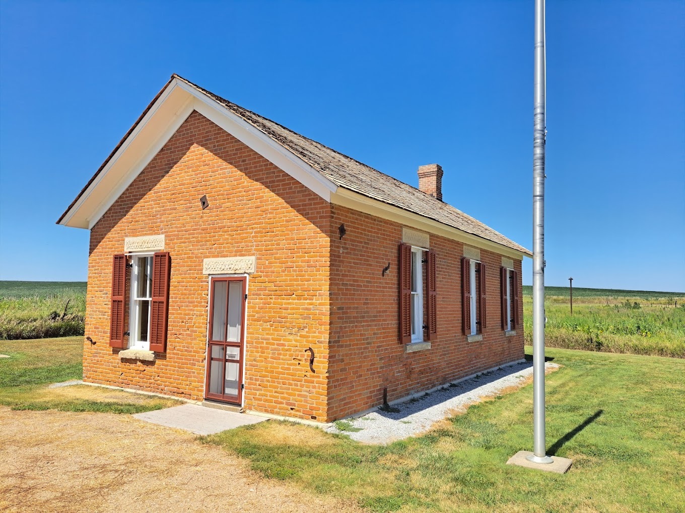 A small one room house with many windows, placed in an empty field, only a flag pole is next to the building.