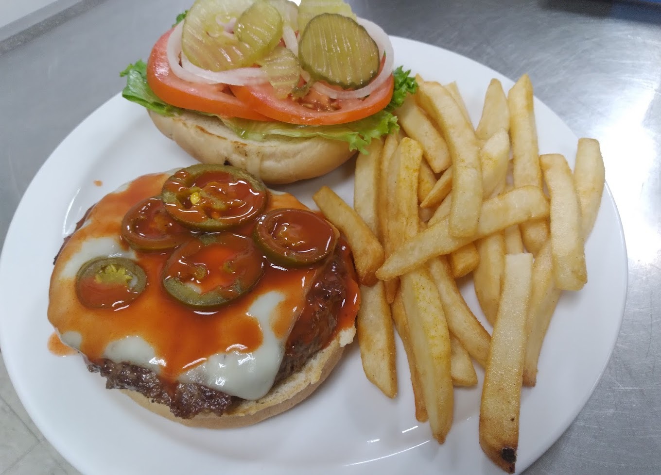 A white plate with a burger with hot sauce, peppers, pickles, tomatoes, and lettuce with a side of fries.