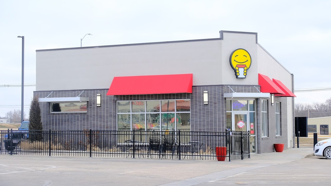A small grey and red accented building, with a large yellow smiley face with a coffee above the door of the establishment.