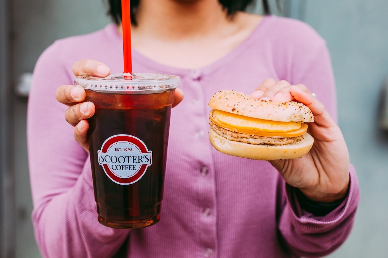 A woman holding an iced coffee beverage in one hand and a breakfast sandwich in the other.