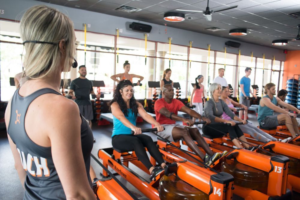 Landscape view of inside of a gym workout room, a lady with a headset is training a group of people working out on equipment.