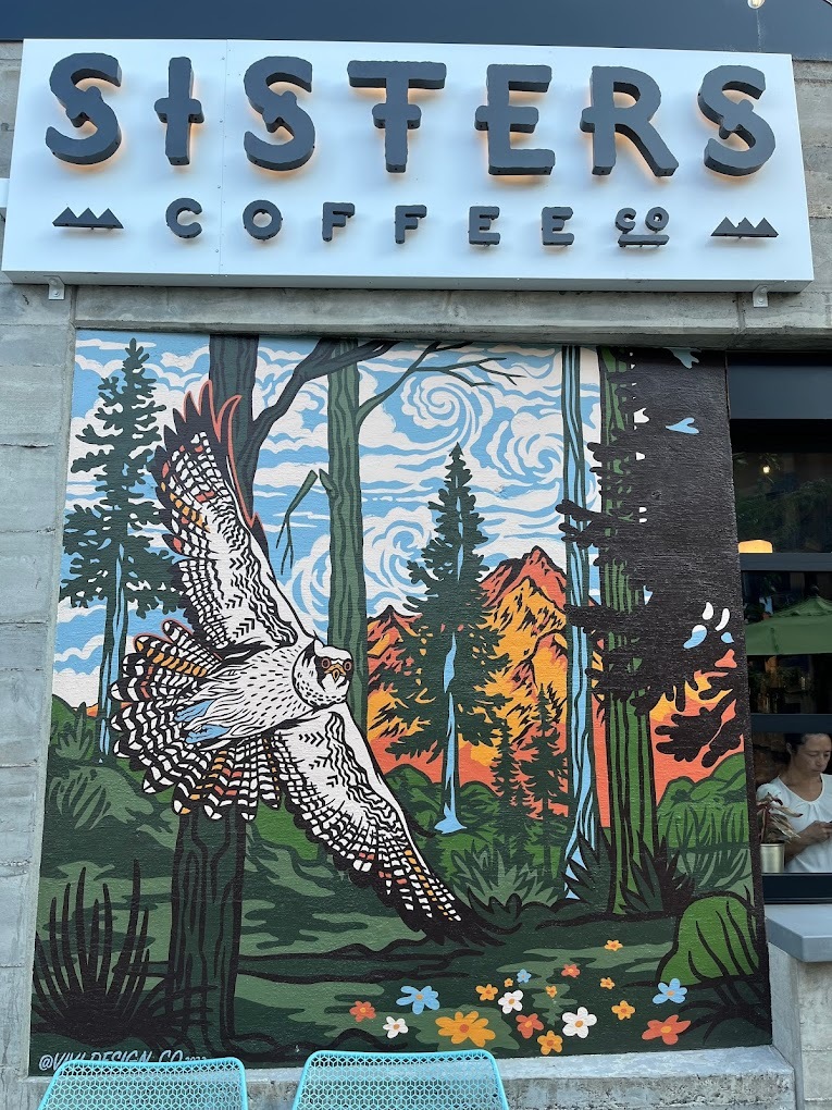 Storefront picture of a decorated window display, depicting an bird flying through a nature scene. Sign at the top of the storefront reads "Sisters Coffee"
