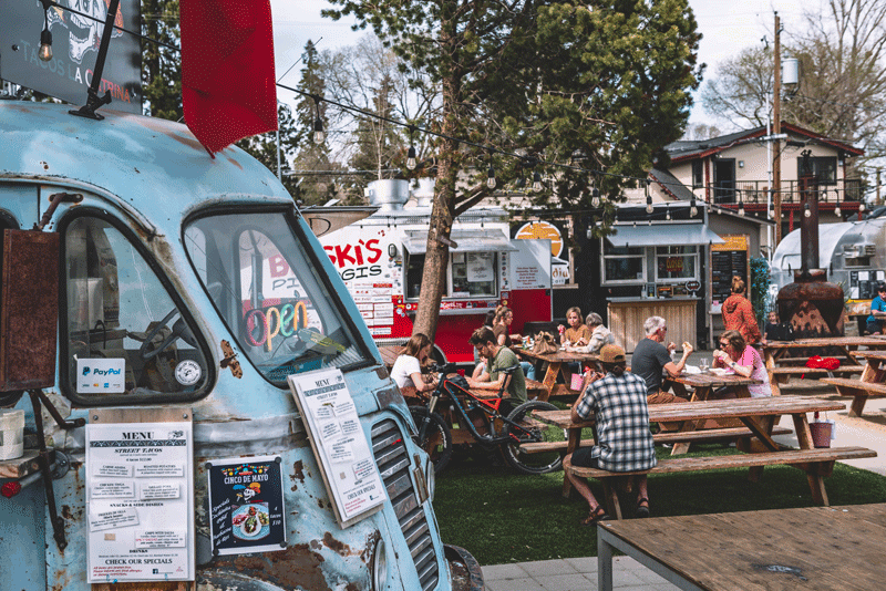 A photo of a patrons sitting on picnic tables surrounded by various food trucks.