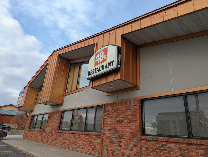Exterior view of a building with the lower half being bricks and several windows. The upper part has vertical slats, and part of the building extends out. In the middle is a grey sign that features a red logo.
