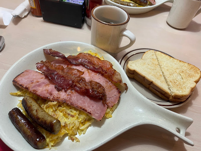 Grey, circular plate with a handle. It has eggs, ham, bacon, and sausage. Beside that is a smaller plate with sliced bread. There is a cup of coffee as well.