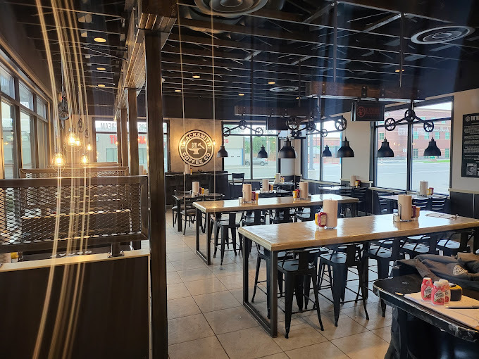 Interior view of a building with light floors, a dark ceiling, rectangular tables with bar stool seating, and hanging lights. The left side of the image has a light flare.