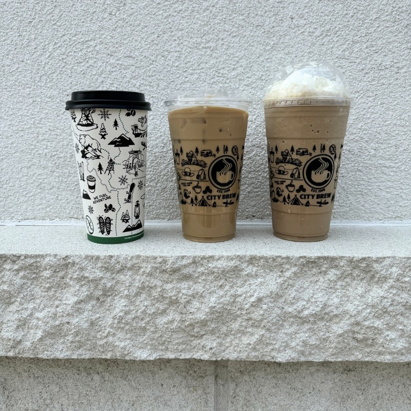Three drinks lined up on a grey, flat, rock structure. The left-most cup is a hot coffee to-go cup, next is an iced coffee, and third is an iced coffee with cool whip. All cups feature outdoorsy doodles.