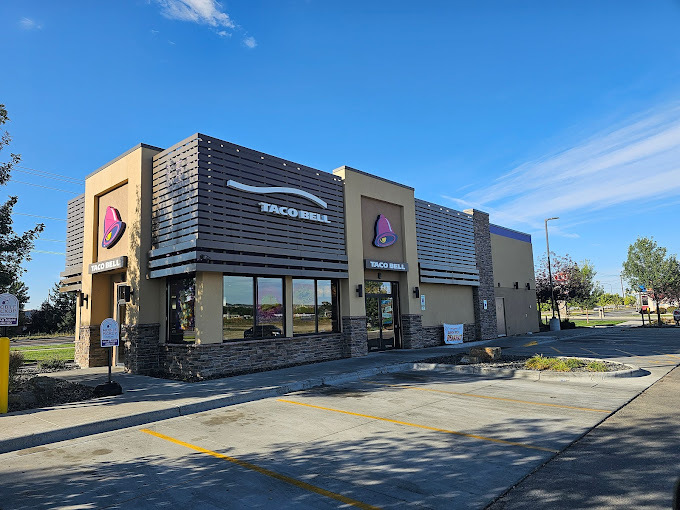 Exterior corner view of a brown building with a brick base and alternating slatted structures above the windows. There are two logo signs on the building, which is a red bell.