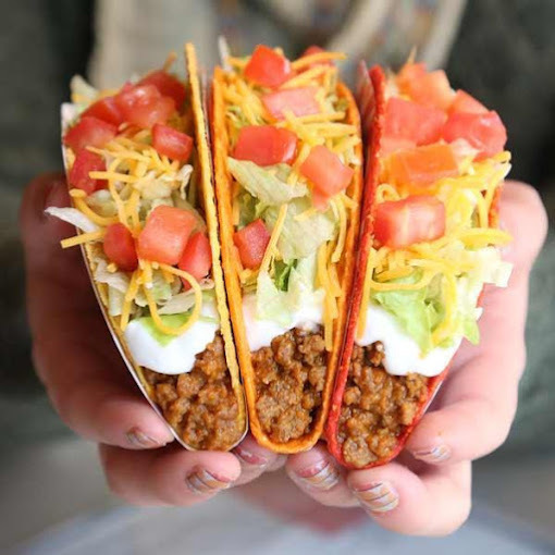 Close view of a person holding three tacos together, presented towards the camera. It features meat, sauce, lettuce, tomatoes, and cheese.