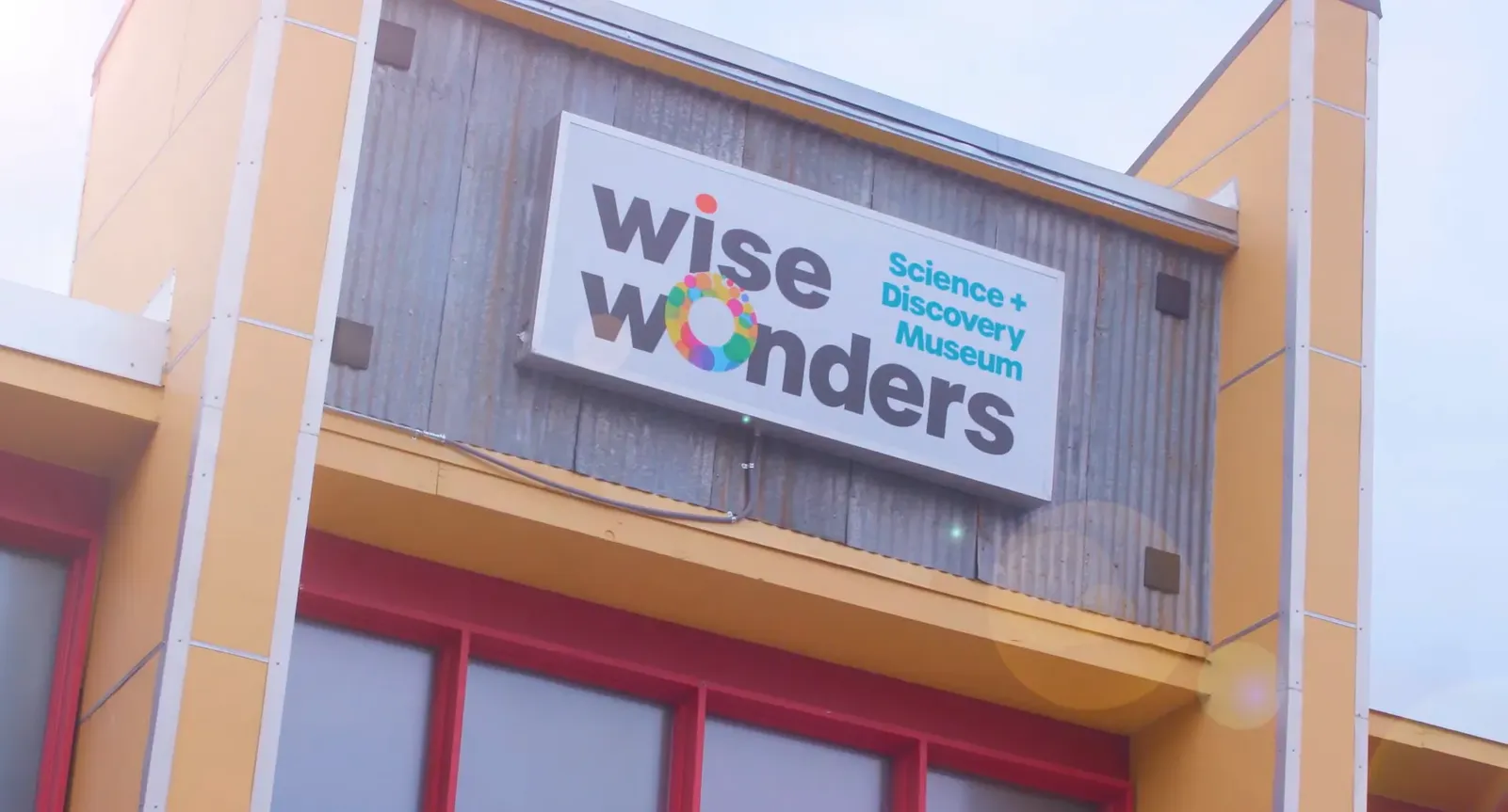 Close view of the sign above the building door entrance. The building is orange and red, with the coloring behind the sign being a grey wood. The logo is black lettering with colorful accents, it reads "Wise Wonders."