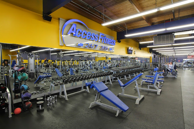 Weight rack and various other blue gym equipment lined up in front of a long wall mirror. The wall behind the mirror is yellow, and the at the top middle of the wall says "Access Fitness" in blue letters.