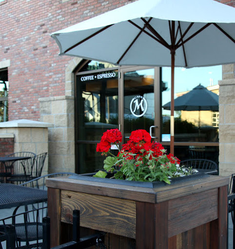 Exterior view of a brick building. The door entrance is arched and has a circular "m" logo in the middle. In front of that, is a table with red flowers and a white umbrella.