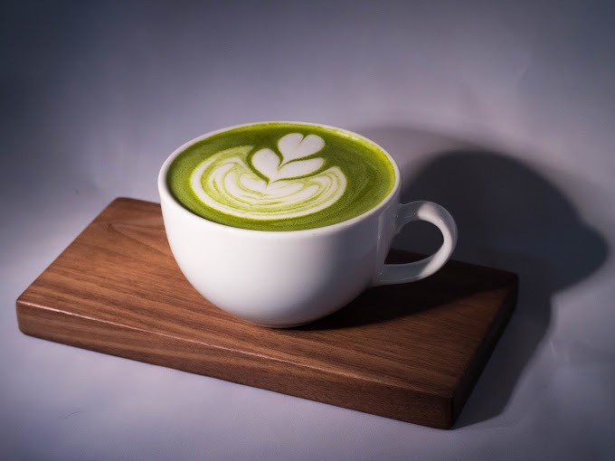 Hot green-colored drink with heart shaped foam art. The white cup sits on top of a small wooden board.