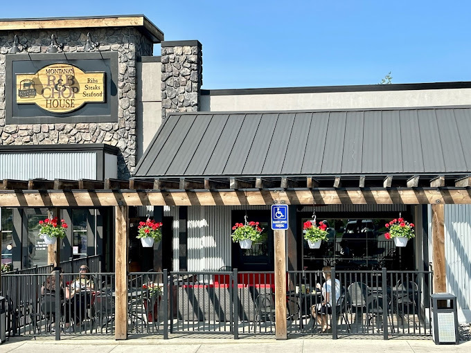 Front exterior view of a light colored building with circular rock accents and a dark colored sun shade. Across the front entrance is a slatted roof with square wooden columns. There is a black fence as well, enclosing a dining area.