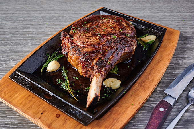 Saucy meat with a large bone on a black rectangular plate, which is on top of a wooden board. Off to the side are a knife and fork.