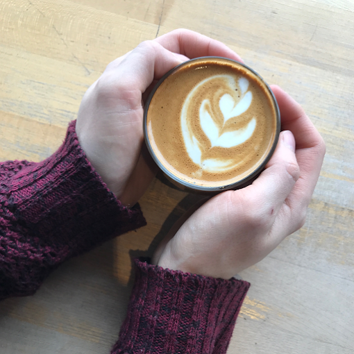 Latte heart-shaped art. Someone holds the sides of the mug with their hands. She is wearing a red long sleeve sweater.