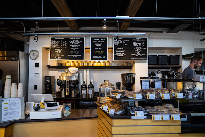 Interior view of a counter area with a  register and menu, as well as coffee making area. Next to the register, there is station of baked goods.