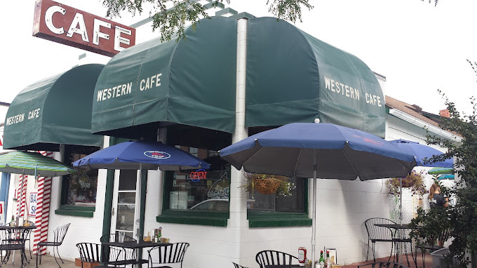 Small white building with puffy green sun shades that say "Western Cafe." There are several tables with blue umbrellas.