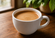 White ceramic mug filled with creamy coffee on a wooden table with a softly blurred natural background.