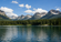 Calm mountain lake bordered by dense evergreen forest and rugged, snow-dusted peaks beneath a bright blue sky with white clouds.