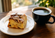 Slice of cinnamon coffee cake dusted with powdered sugar beside a black mug of coffee on a wooden table near a softly lit window.
