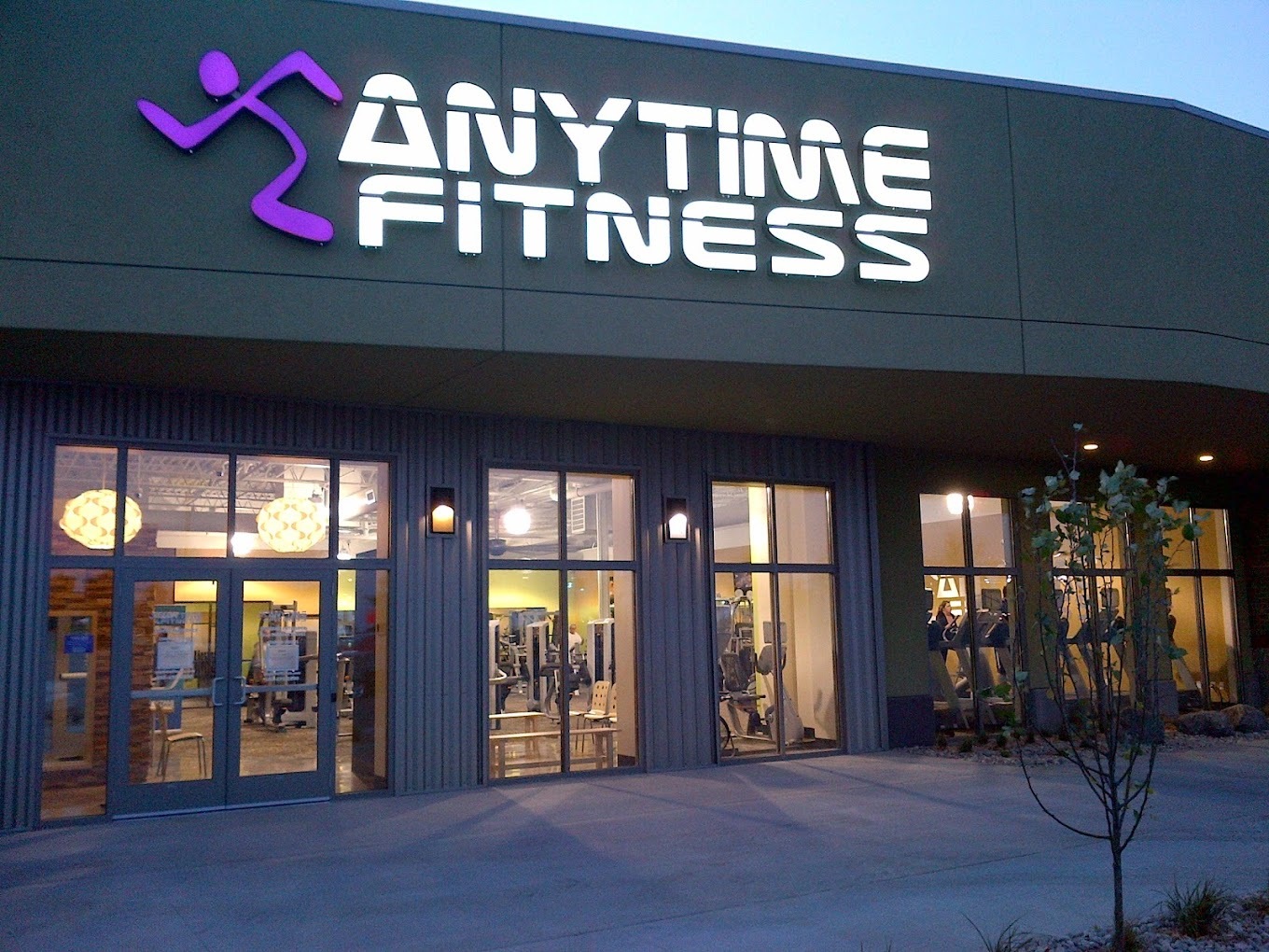 An outside view of an gym with lots of windows and a large lighted sign above the doors that reads Anytime Fitness in white lettering with a purple stick figure logo.