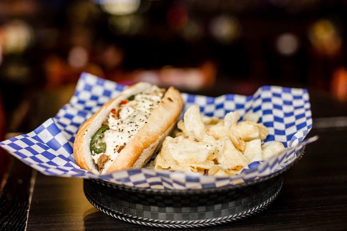 A serving tray with blue checkered paper consisting of a hot dog with toppings and seasoning and served with a side of kettle chips.