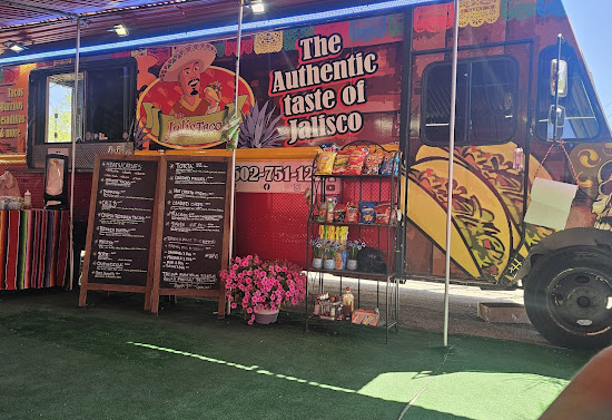 Taco food truck with two menu signs, flowers, and a rack of chips in front of it. These items are on top of a grassy area.