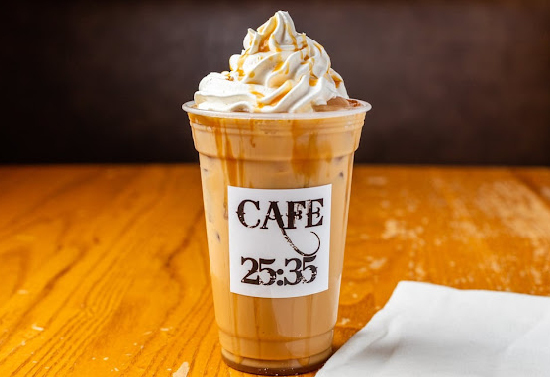 Iced coffee drink with whipped cream and caramel drizzle. The cup has a "Cafe 25:35" sticker. A napkin is on the right side and these items reside on a wooden counter.