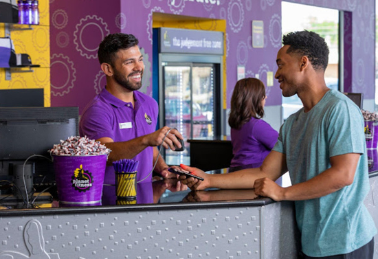 A bearded man in a purple polo scans the phone of a man in a blue shirt. There is a counter with a computer between them. On the counter there are two containers of popcorn and pens. In the background, a woman in a purple shirt faces away from the camera.