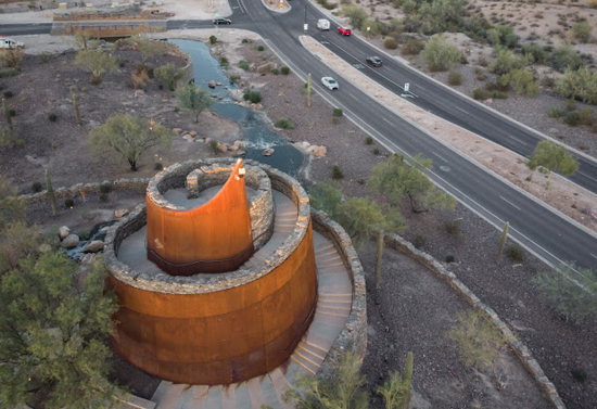 Spiral orange building with stairs and rock decoration is on the bottom left of the image. The upper left of the image has a bridge going over a small river and the right of the image has a highway road.