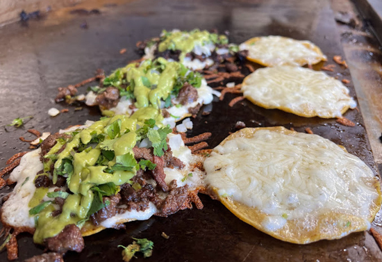 A set of six egg patties are on a brown stone grill. The three on the left have meat and green sauce whereas the three on the right do not.
