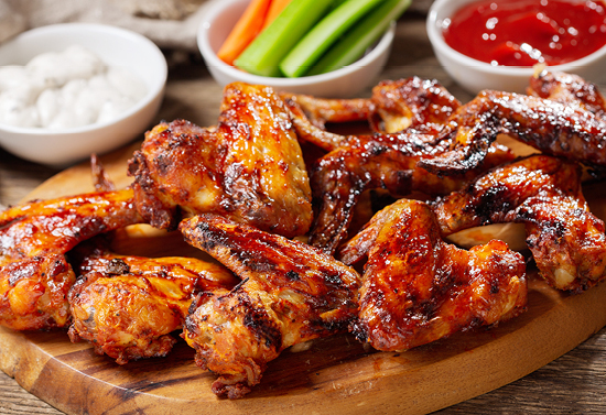 Fresh cooked wings gathered on a wooden plate. Three different ups of sauce, veggies, and ketchup are in the background.