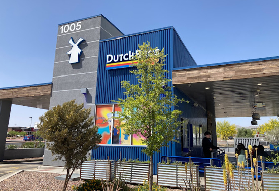 Upward view of the side of the Dutch brothers building. It's gray and navy with patio covers on either side. Small trees are towards the front of the image.