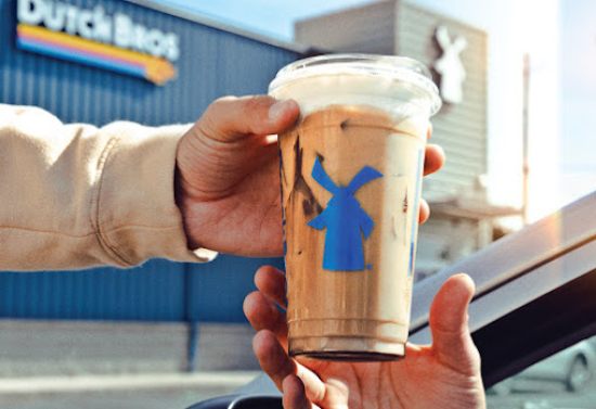 Close view of a man in a beige sweater handing an iced coffee to a man sitting in his car. The Dutch brothers building is seen in the background.