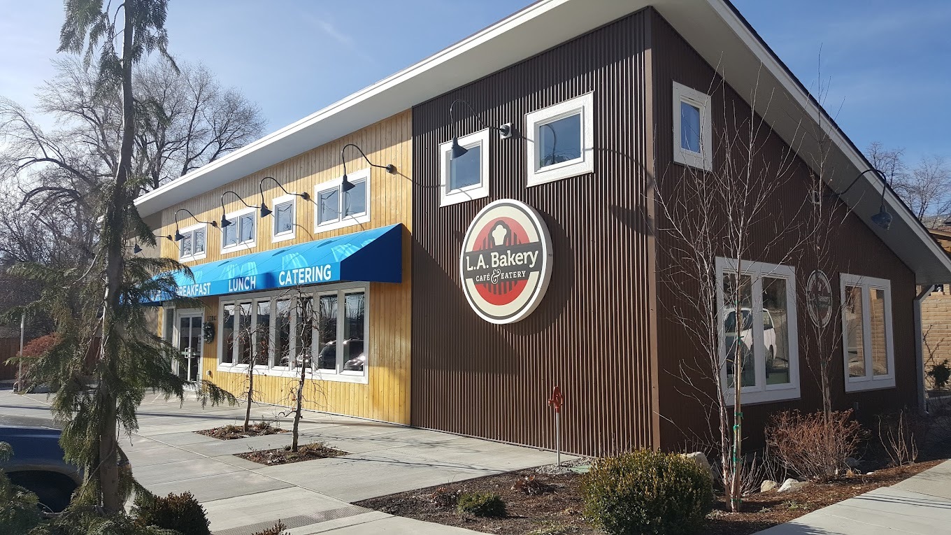 an outside view of a pastry shop that is a two tone yellow and brown building accented with a blue awning and a large round sign that reads "LA Bakery"