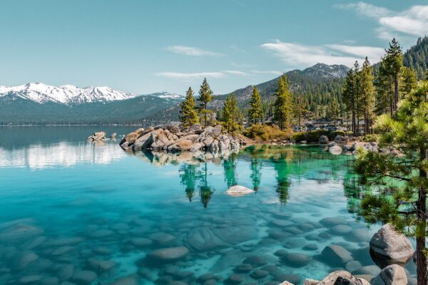 A scenic landscape view of the lakes shore. Mountains with snow caps can be seen in the background. The lake is a dark clear blue color.