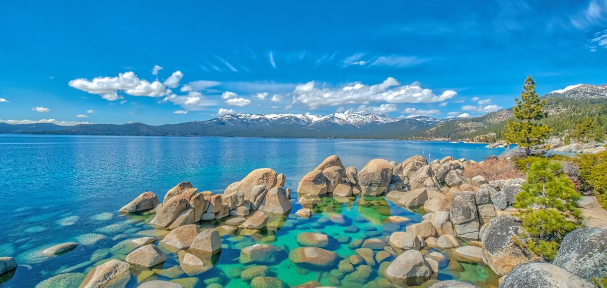 Scenic landscape view of a blue sky and a lake with large rocks and trees in the shore. The lake's water is a deep blue and lighter blue at the shore.