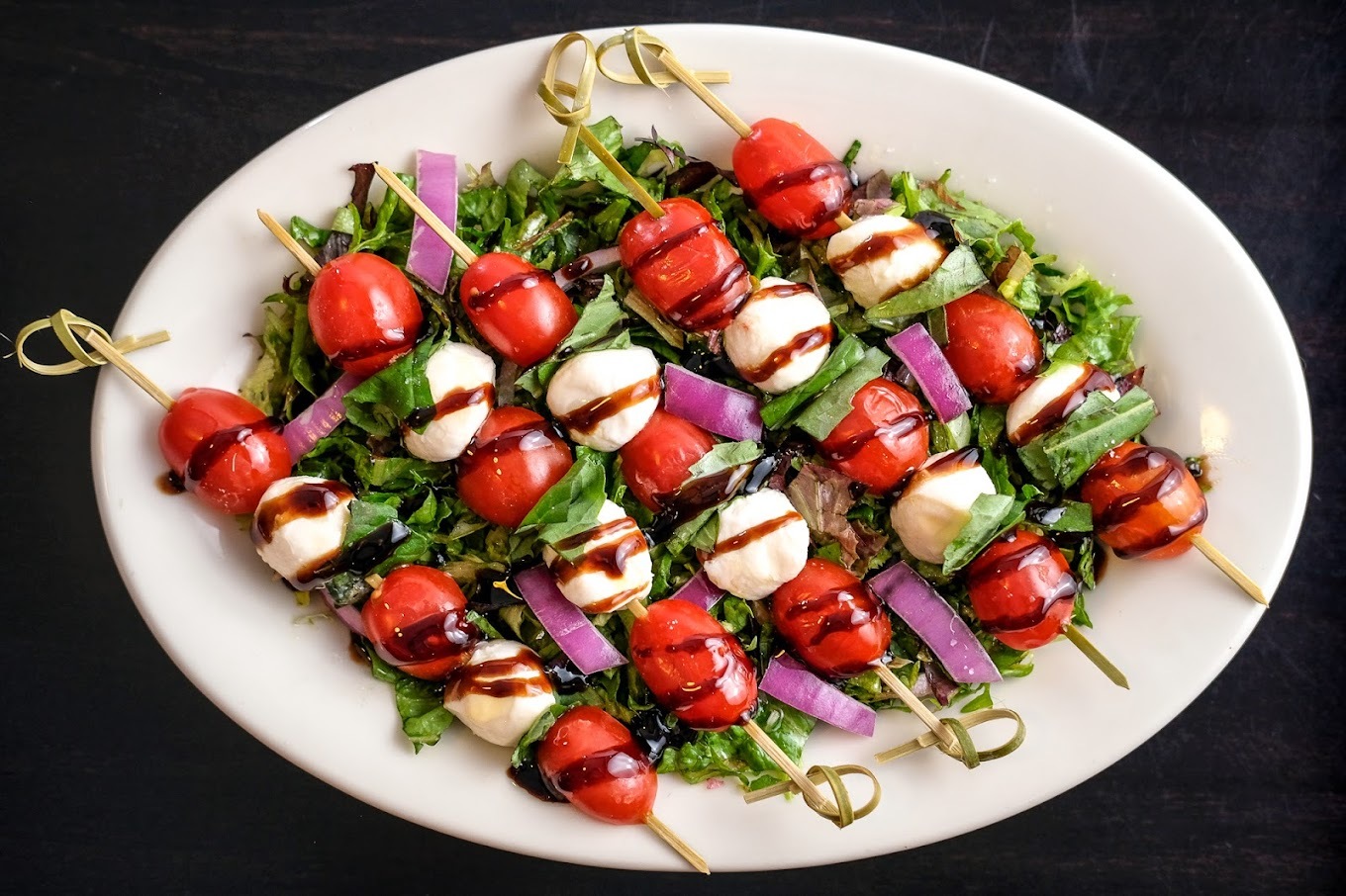 A white plate with a salad base, setting atop are skewers of tomato and mozzarella drizzled with balsamic vinaigrette.