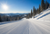 Groomed ski slope winding through snow-covered pine trees with mountain views under a clear winter sky.