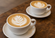 Two white ceramic lattes with leaf latte art, one in sharp focus in the foreground and a second softly blurred in the background.