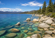 Crystal-clear mountain lake with smooth granite boulders, evergreen trees, and distant snow-capped peaks beneath a bright blue sky.